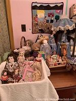 Front view of multiple dolls grouped on a table and basket with cloth covers, showing various porcelain and cloth dolls and some doll lamps with detailed lace lampshades.