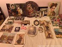 Wide shot of small framed art pieces and plaques laid out on a surface, including portraits and scenes with children and flowers, multiple frame shapes and sizes.