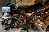 Overview of many vintage tools and hardware items laid out, including a hard hat, silver metal toolbox, black oil can, and smaller hand tools.