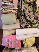 Stack of assorted folded fabrics, lace trims in pastel pink and yellow, heavier woven fabrics, and wool blankets showing texture and some wear.