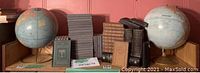 Wide view showing two globes on either end and an assortment of vintage and antique books in the middle on a wooden surface, with a pink wall background.
