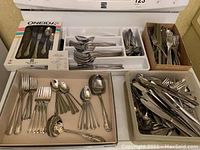 Multiple flatware pieces displayed in trays and boxes showing forks, spoons, serving utensils, and boxed Oneida set in unopened package.