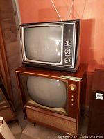 Two vintage TVs stacked with visible control knobs and screens, one with wood finish and fabric speaker grille, the other black and silver portable TV.