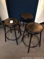 Three identical National Public Seating metal stools with black legs and dark brown wooden tops shown from an angle, arranged in a triangular formation on carpet.