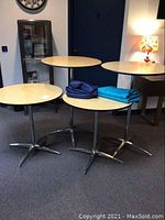 Four round tables with light wood laminate tops, metal bases, and five round tablecloths folded on top, displayed in room setting