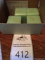 Four large square votive candles in honey dew color inside an open box placed on a wooden table, with a paper auction tag partly visible.