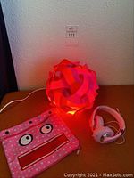 Photo showing the glowing pink and red puzzle light plugged in with pencil case and headphones beside it