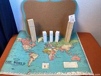 Photo showing scalloped cork bulletin board with teal frame standing behind a large vintage world map lying flat, with multiple bundles of natural and white wooden craft clothes pins arranged on the map.