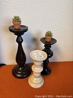 Photo of three pillar candle holders on a flat surface against a wall. Two holders are black and one is white, each topped with a small succulent candle in a miniature terracotta pot.