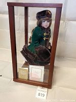 Full view of Anna Katharina doll seated inside wooden framed glass display case showing green velvet coat with dark brown faux fur trim and included tags and literature in the case.