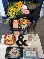 Photo shows assorted faux flowers in yellow, pink, and white, black decorative ampersand, glass vases, Ed Hardy handbag with colorful graphics, perfume tubes, and decorative signs.