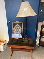 Photo showing a wood piano bench with black velvet seat, accompanying large floor lamp with cream lampshade and decorative black and gold stand, and wooden serving tray with lace design under glass top placed on bench.
