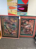 Two large framed mixed media abstract art pieces, one pink frame and one teal green frame, both with hanging wire on back. Artwork is raised and textural with torn paper and string elements.