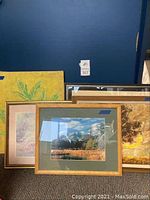 Five artwork pieces including framed and matted landscape print with mountain scene in wooden frame with glass; several other artworks behind it showing various paintings and framed prints.