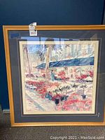 Frontal photo of the colorful painting showing outdoor market scene with flower pots and plants, framed in wood with glass and cream/navy matting