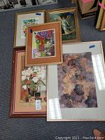 Five framed artworks laid on floor, showing varied sizes and frame colors including rose gold, gold, brown, and natural wood. Visible damaged corners and frames.
