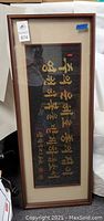 Full front view of the framed Asian calligraphy artwork showing gold characters on a black textured background within a brown wooden frame and beige matting.
