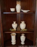 Photo showing three shelves with seven white Lenox porcelain items and two clusters of plastic grapes used as decoration. Items include various vases and dishes.