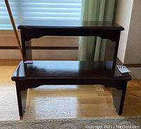 Two-step wooden library steps shown in front of window with blinds and curtain. Dark finish, smooth top and bottom steps, with visible scratches and wear on surface.