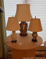 Three lamps on round table: one Asian theme lamp with decorative base and rectangular beige shade, and two smaller floral ceramic lamps with beige shades.