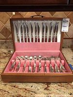 Wooden chest with pink felt interior open to display 14 knives in lid and assorted forks and spoons in base on granite countertop.