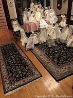 Group display of multiple porcelain bridal dolls in various white and ivory satin and lace wedding gowns, arranged on furniture and carpets in a room.