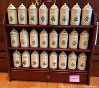 Front view of wooden spice rack filled with 24 Lenox Spice Garden ceramic spice jars arranged in three rows with drawer below.