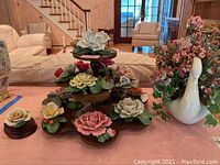 Full view of thirteen porcelain flower figurines placed on three-tier dark wooden stand and smaller stand, alongside a white ceramic swan vase holding artificial floral bouquet.