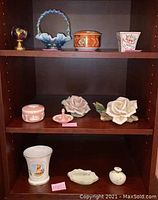 Shelf with assortment of decorative and collectible porcelain and ceramic items including Wedgwood box and ring holder, Capodimonte roses, orange trinket box, and glass decor pieces.