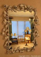Full view of the rectangular wall mirror with white metal frame decorated with raised floral and leaf patterns, reflecting a living room interior.