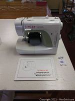 Front-left view of white Singer Simple model 3116 sewing machine with green accent and instruction manual opened on table.