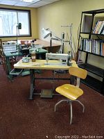 Wide view showing sewing machine mounted on table, swivel chair, lamp, and surrounding workshop area