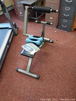 Side angle of Body by Jake workout bench showing padded seat and leg support structure, placed on carpet in room near filing cabinets and treadmill.