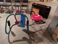 Two exercise machines on basement floor: red DK abdominal sit-up exerciser with foldable frame and arm supports, and blue/pink thigh exerciser with resistance band and foam padded handles.