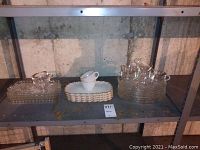 Overall view of shelves holding 4 glass trays in stack, 2 china cups resting in baskets, 2 glass cups, and stacks of glass plates.
