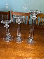 Frontal view of three clear lead crystal candle holders placed on a wooden table, showing varying heights and design details.