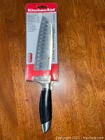 Front view of KitchenAid 7-inch Santoku kitchen knife in clear packaging on wooden table.