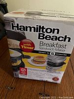 Front and side views of the Hamilton Beach Breakfast Sandwich Maker box, showing brand, timer feature, and sandwich image.