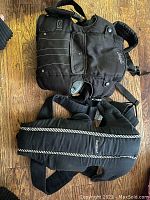 Two forward-facing baby carriers laid flat on a wooden floor, one black Baby Bjorn carrier with padded straps and white trim, and one black Snugli carrier.