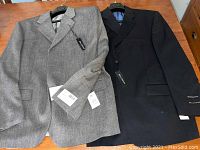 Two men's suit jackets laid flat on table, showing front sides. Black and grey colors, tags attached.