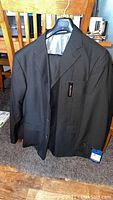 Photo of black pinstripe men's suit jacket and trousers hanging on chair, showing front view and brand tag.