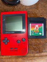Front view of red Game Boy Pocket handheld console and Babe game cartridge side by side on wooden surface.