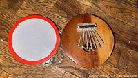 Photo of a small tambourine with red rim and a coconut shell calimba thumb piano with six metal tines on wooden floor.