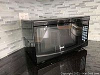 Black RCA microwave oven placed on a black countertop against a tiled backsplash, front and side view showing keypad and vent