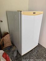 Side and door view of the white Danby Premiere bar fridge with beige trim sitting on granite floor next to bags and a lamp.