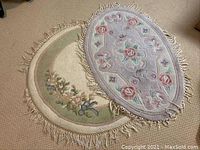 Two wool rugs overlapping; one oval with pink and lavender floral design and one round with beige and green floral design, both with fringed edges.