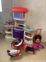Photo showing a wide view of the Little Tikes kitchen playset, Mickey Mouse rolling bag, Captain America shield, pink bowling pins, and other toys on carpeted floor.