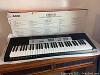 Image of the Casio CTK-1500 keyboard on a wooden dresser with its box behind it displaying multilingual model information.
