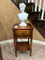 Frontal view of wooden side table with drawer and white plastic bust on top, showing the bust intact from the front and the drawer's ornate handle.