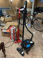 Two vacuum cleaners shown with a bicycle and boxes in the background, one red stick vacuum with a cord and one black Bissell canister vacuum with hose and floor head.